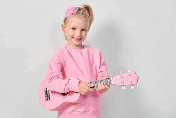 可愛いですCaucasian女の子4 5年古いですピンクスウェットシャツあります遊んでいますザUkulelに対してグレースタジオ背景 — ストック写真