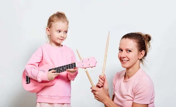 Yong mother and her pretty daughter playing on musical instruments, neutral gray background. Spending funny time together holding ukulele and wood drumsticks.