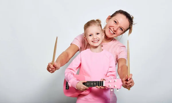 Yong mother and her pretty daughter playing on musical instruments, neutral gray background. Spending funny time together holding ukulele and wood drumsticks.