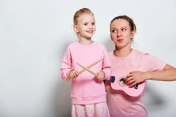 Yong mother and her pretty daughter playing on musical instruments, neutral gray background. Spending funny time together holding ukulele and wood drumsticks.