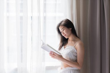 Asian women while reading book near window with shear curtain / 