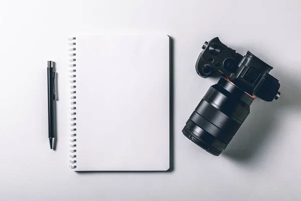 Tavolo da ufficio moderno bianco con penna e macchina fotografica digitale mirrorless. Pagina bianca del taccuino per inserire il testo al centro. Vista dall'alto, piatta . — Foto Stock