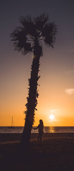 Беременная женщина прикасается к животу и смотрит на закат, рядом с пальмой, в Альмеримаре, Альмерия, Испания
