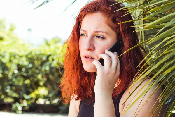 Primer plano de chica seria con pelirroja hablando en el teléfono móvil mientras está de pie al aire libre — Foto de Stock