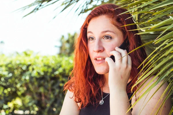 Primer plano de chica sorpresa con pelirroja hablando en el teléfono móvil mientras está de pie al aire libre — Foto de Stock
