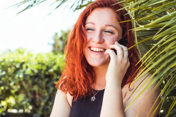 Primer plano de chica atractiva sonriente con pelirroja hablando en el teléfono móvil mientras está de pie al aire libre — Foto de Stock