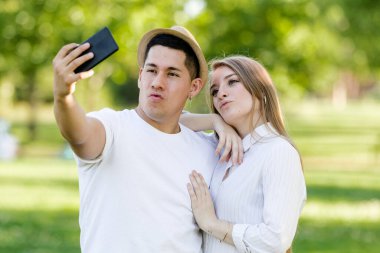 Parkta cep telefonlarından selfie çeken genç bir çift. Latin erkek ve beyaz kadın.