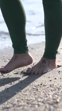 İsimsiz bir kadının, kumlu sahilde, gün ışığında, yeşil tozluklarla çıplak ayakla yürümesi.