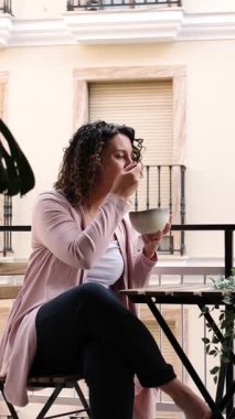 Balkon masasında oturan genç bir kadın, sakin bir sabah yemeğinin keyfini çıkarıyor, sakin bir tavırla, sağlıklı bir kahvaltının tadını çıkarırken kentsel çevreye bakıyor. Dikey görüntüler