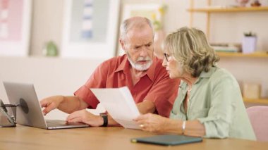 Yaşlı çift evrakları gözden geçirir ve dizüstü bilgisayar kullanır, konforlu ev ortamlarında mali veya diğer önemli konuları tartışır