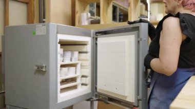 Potter eldiven takıyor, çömlek atölyesinde elektrikli fırının içindeki raflara bardak koyuyor.