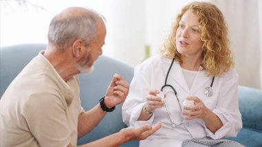 Elinde ilaç şişesi olan doktor, evde bakım ziyareti sırasında kanepede rahat bir şekilde oturan yaşlı bir adamla reçete detaylarını tartışıyor.