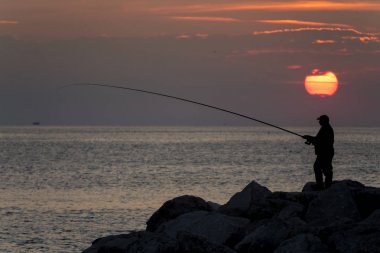 gün batımında kaya üzerinde duran denizde adam balıkçılık