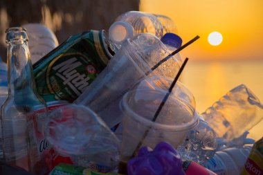 Faliraki, Rodos, Yunanistan - 11 Temmuz 2019: Çöp, plastik şişeler, kokteyl tüpleri ve içki tenekeleri sahilde ki çöp kutusunda gün doğumu veya gün batımının arka planında gökyüzü ve palmiye ağaçlarına karşı se