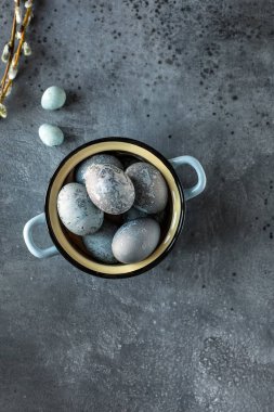 Gri taş dokulu arka plan üzerinde mavi bir vintage tencerede Boyalı Paskalya yumurtaları. Arka planda iki bıldırcın yumurtası ve söğüt dalları. Üstte görüntü.