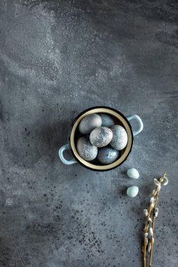 Gri taş dokulu arka plan üzerinde mavi bir vintage tencerede Boyalı Paskalya yumurtaları. Arka planda iki bıldırcın yumurtası ve söğüt dalları. Üstte görüntü.