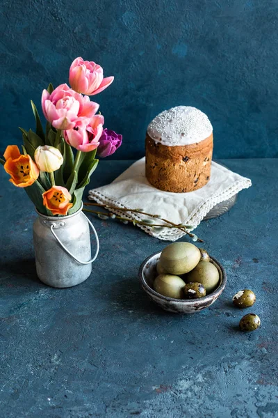 Easter composition. On a blue textured stone background a bouquet of tulips in a vintage bucket, painted Easter eggs, Easter cake and willow. Close up.