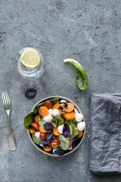 Fesleğenli yeşil mozzarella salatası, domates kiraz, limonlu bir bardak su. gri beton dokusal arka plan. Üstte görüntü.