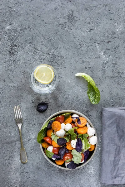 Fesleğenli yeşil mozzarella salatası, domates kiraz, limonlu bir bardak su. gri beton dokusal arka plan. Üstte görüntü.