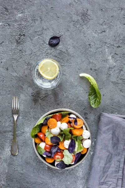 Fesleğenli yeşil mozzarella salatası, domates kiraz, limonlu bir bardak su. gri beton dokusal arka plan. Üstte görüntü.