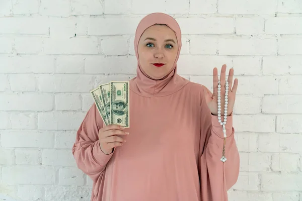 Λευκή γυναίκα με μπλε μάτια σε ροζ χιτζάμπ κρατώντας ένα κομπολόι και δολάρια σε λευκό φόντο. — Φωτογραφία Αρχείου