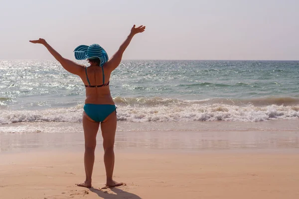 Happy elderly woman in a hat with raised hands on a background of the sea at sunset. Concept of sunny and happy summer.