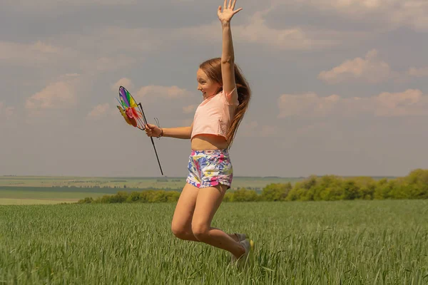 Menina feliz com cabelos longos segurando um moinho de vento colorido e salta . — Fotografia de Stock