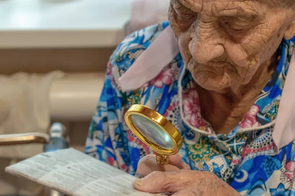 Mycket gammal kvinna med förstoringsglas försöker läsa från en tidning. Mormor 90 år gammal läser vid bordet med förstoringsglas . — Stockfoto