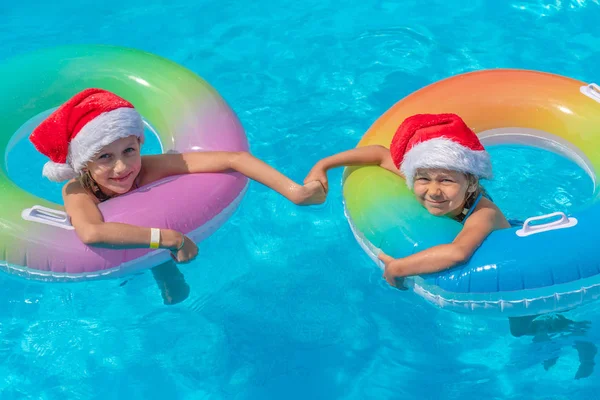 Две девушки в шляпах Санта-Клауса купаются в голубом бассейне в яркий солнечный день и улыбаются. Концепция счастливого нового года и Рождества — стоковое фото