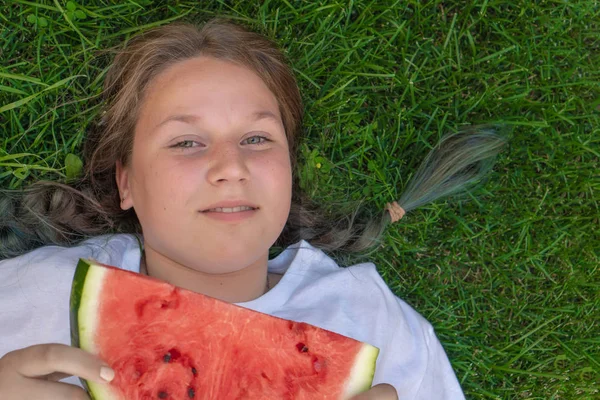 Portrait of a fat girl of 13 years old who lies on the grass with a watermelon in her hands