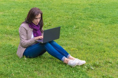 Yeşil çim üzerinde oturan parkta dizüstü bilgisayar kullanan çekici kadın