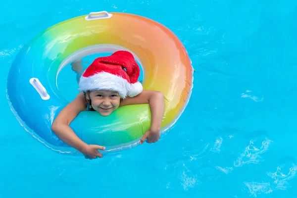 Ребенок в шляпе Санта-Клауса плавает в голубом бассейне в яркий солнечный день и улыбается. Концепция счастливого нового года и Рождества — стоковое фото