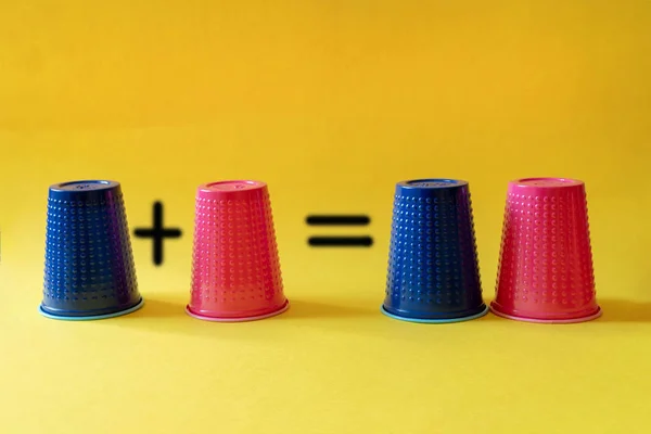 Math exercise: one plus one equals two. Multi-colored blue and pink plastic cups on a yellow background