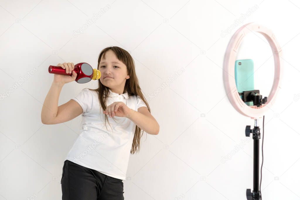 A teenage girl with a microphone in her hand sings and shoots a video. Selfies. The phone is mounted on a tripod and the ring lamp shines. Emotions Girl blogger