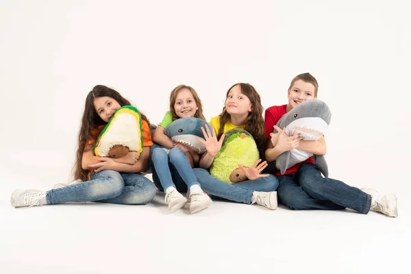 Un grupo de niños en ropa brillante con sus juguetes suaves favoritos en sus manos sobre un fondo blanco. Foto del estudio. Bebé moderno tendencia suave juguete aguacate y tiburón — Foto de Stock