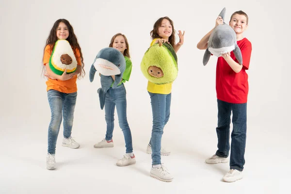 Sekelompok anak-anak dengan pakaian cerah dengan mainan lembut favorit mereka di tangan mereka dengan latar belakang putih. Foto studio. Baby modern tren lembut mainan alpukat dan hiu — Stok Foto