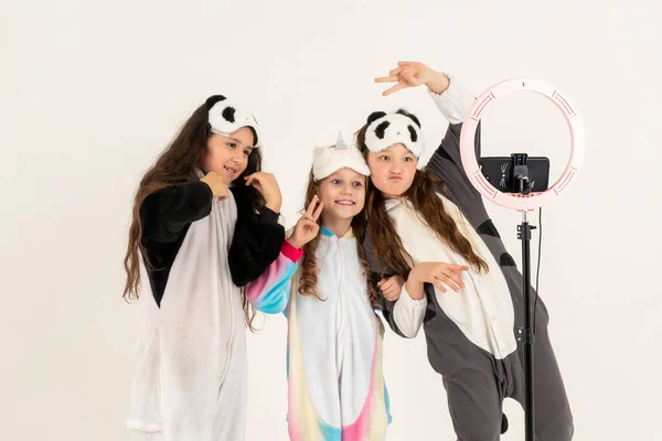 Gadis remaja yang lucu di kigurumi dan topeng tidur tersenyum dan merekam video. Selfie. Ponsel dipasang pada tripod dan lampu cincin bersinar. — Stok Foto