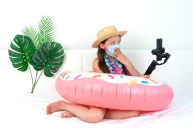 Küçük kız blogcu evde yatakta otururken tatil taklidi yapıyor, video bloğu kaldırıyor. Yüzme çemberinde ve tıbbi maskeli bir çocuk evde kalır ve sosyal ağlar için video çeker. Yurtiçi turizm ve koronavirüs...