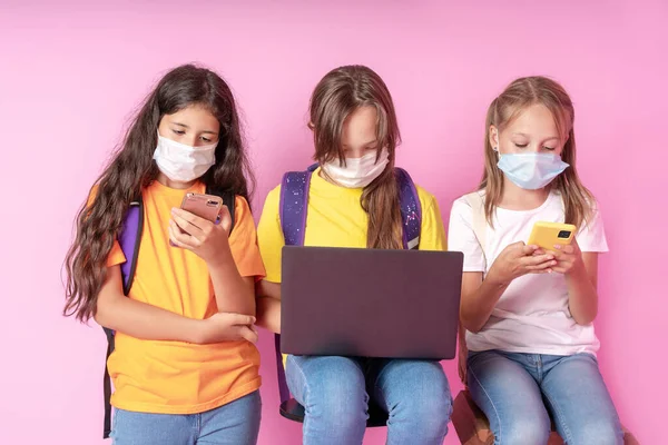 Три школярки в медичних масках тримають смартфони і ноутбук. Концепція дистанційного навчання. Повернутися до школи. Студійне фото . — стокове фото