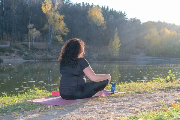 Fat caucasian woman with long curly dark hair dressed in black tshirt and black sportive pants online is doing yoga on purple mat near the lake in the autumn park. The phone shows an online yoga class