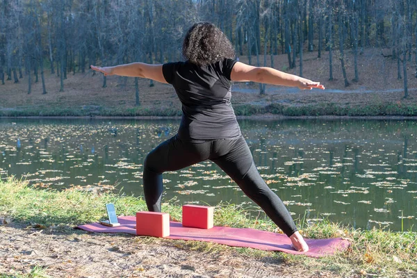 Fat caucasian woman with long curly dark hair dressed in black tshirt and black sportive pants online is doing yoga on purple mat near the lake in the autumn park. The phone shows an online yoga class