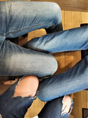 Ahşap bir zeminin arka planında kot pantolon giymiş üç çift bacak. Bazı kotlar yırtılmış..