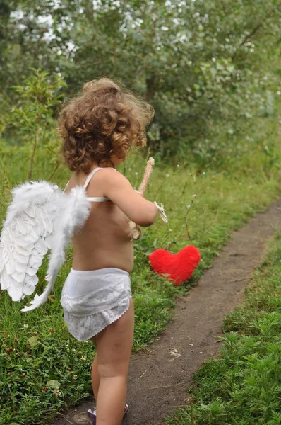 Niño de pelo rizado con alas brota de un arco en el corazón rojo . — Foto de Stock