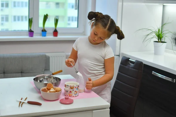 girl pours sugar into a measuring cup with a spoon. Little girl preparing cookies in kitchen at home. Cooking homemade food.