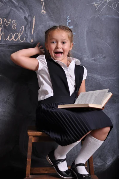 Portret pięknego uśmiechniętego młodego pierwszego równiarka siedzi z książką na tle tablicy. koncepcja szkolna i edukacyjna — Zdjęcie stockowe