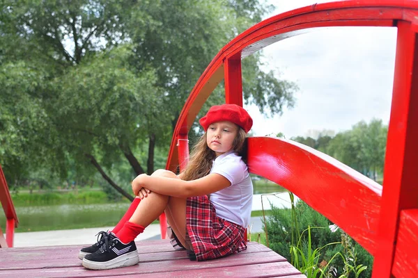 Retrato de una hermosa colegiala en sombrero rojo en el parque de fondo y puente rojo — Foto de Stock