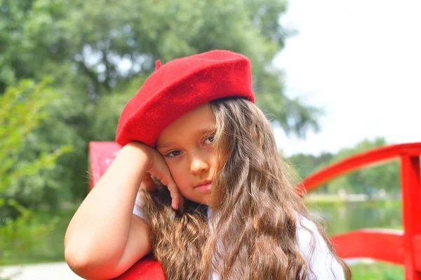 Retrato de la triste joven colegiala en sombrero rojo en el parque de fondo y puente rojo — Foto de Stock