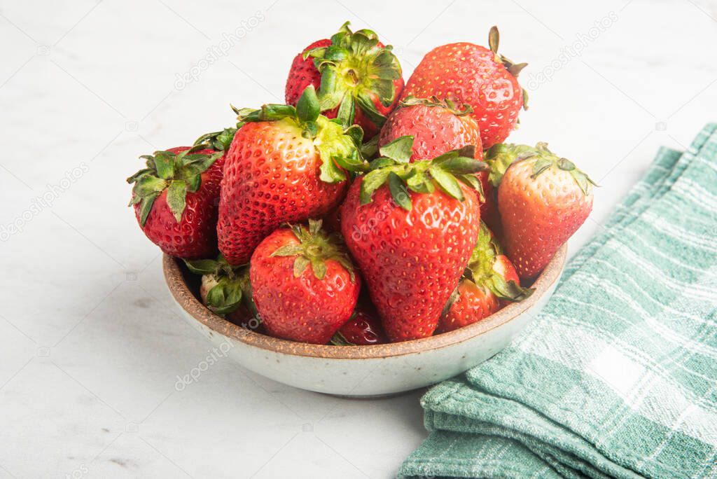 A ceramic bowl of fresh and sweet red strawberries set on a marble countertop with green napkin.