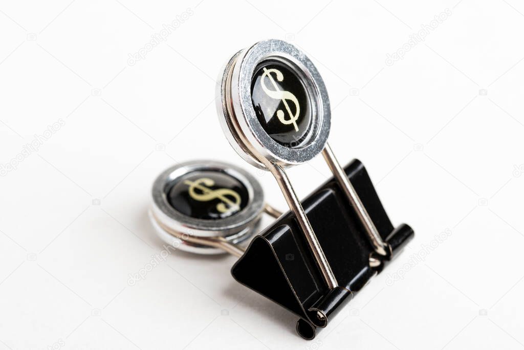 A close-up of a black document clip with circular press handle featuring a dollar sign set on a plain white background.