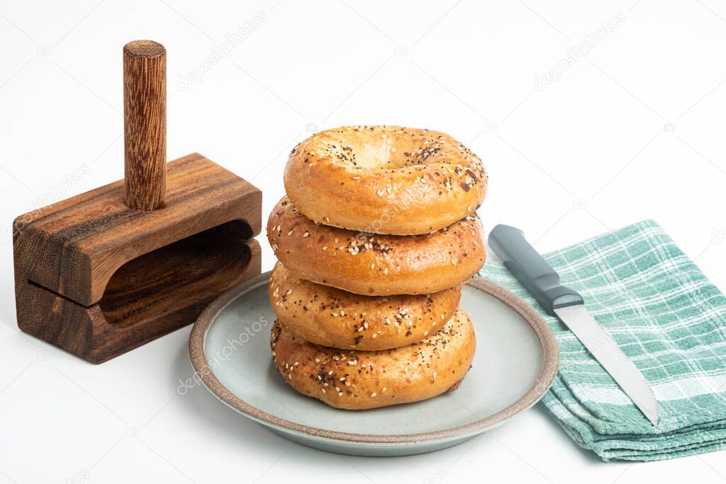 A single tall stack of four freshly baked bagels on a ceramic plate with a cutting wood stand and knife set on a plain white background.